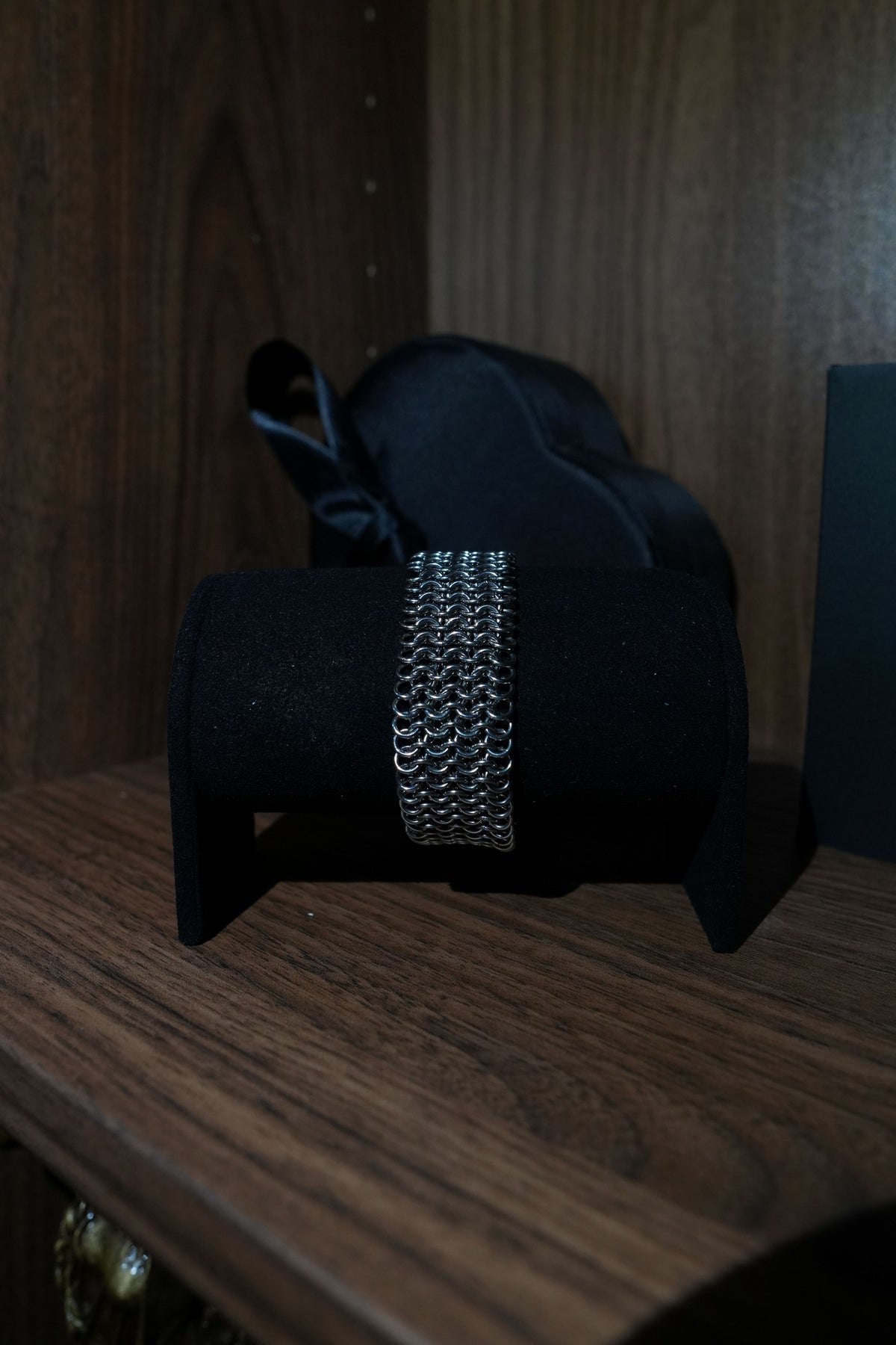 A silver-toned chain-link bracelet displayed on a black velvet jewelry stand, set on a dark wooden surface with a black satin bow in the background.
