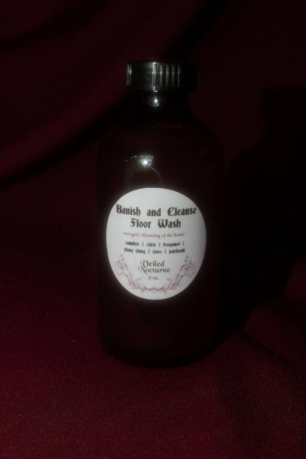 A dark amber glass bottle of 'Banish and Cleanse Floor Wash' by 'Deeded Nectarine' with a white circular label and black cap, set against a dark red fabric background.