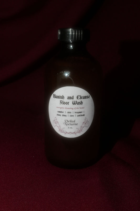 A dark amber glass bottle of 'Banish and Cleanse Floor Wash' by 'Deeded Nectarine' with a white circular label and black cap, set against a dark red fabric background.