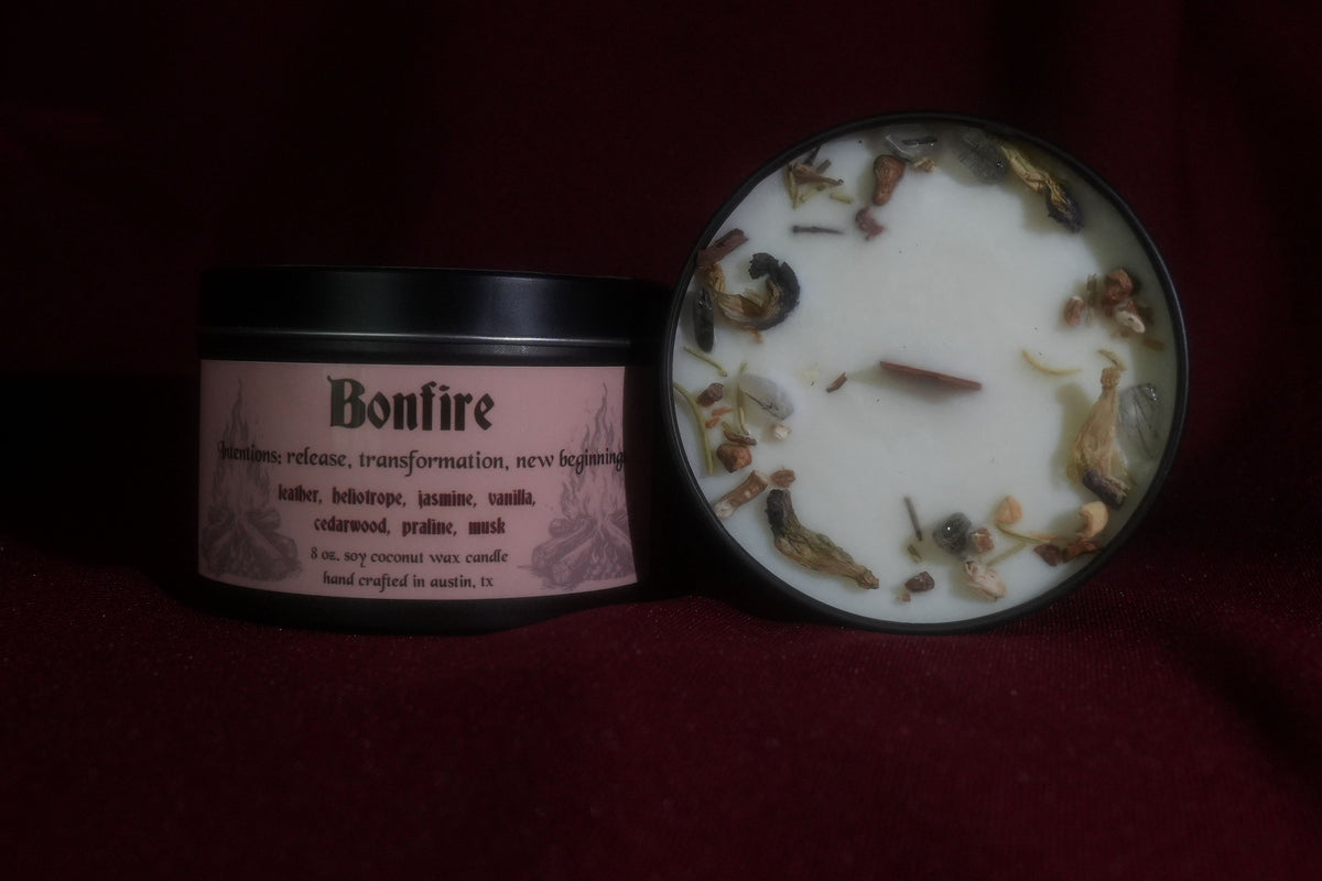 A candle product shown in two pieces on a dark red fabric background: a black-lidded jar with a pink label reading Bonfire, and the open white wax top with dried botanicals scattered around the rim.