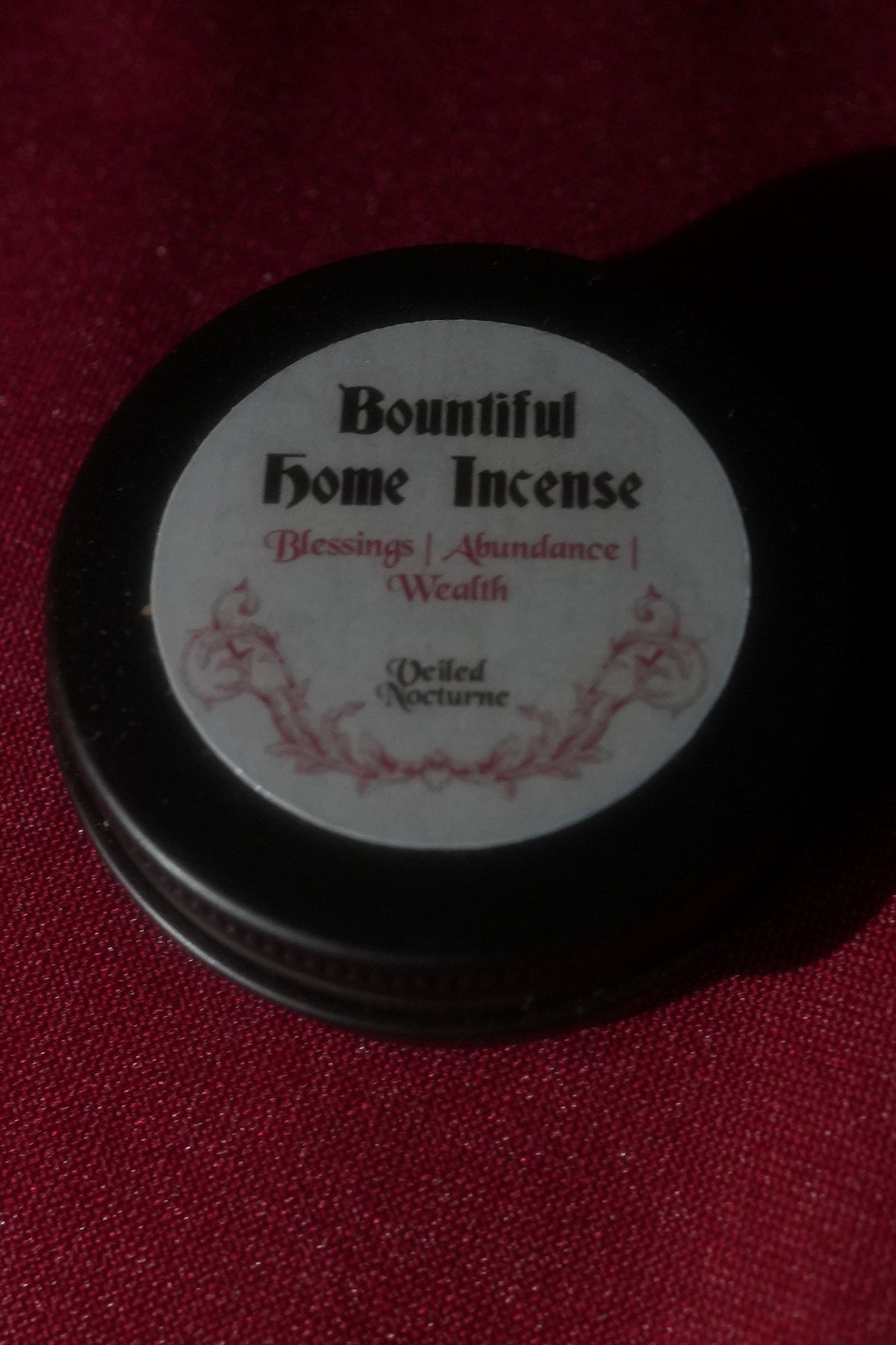 A small round black tin of Bountiful Home Incense with a white label, placed on a dark red textured fabric background.