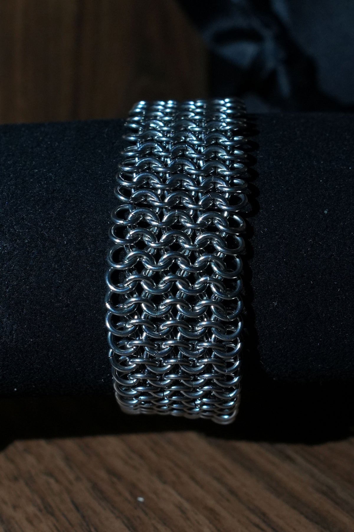 A silver metallic chainmail-style cuff bracelet displayed on a black cylindrical stand, photographed against a dark background with a wooden surface visible at the bottom.