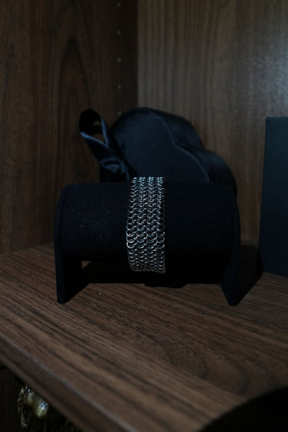 A silver-toned chain-link bracelet displayed on a black velvet jewelry stand, set on a dark wooden surface with a black satin bow in the background.