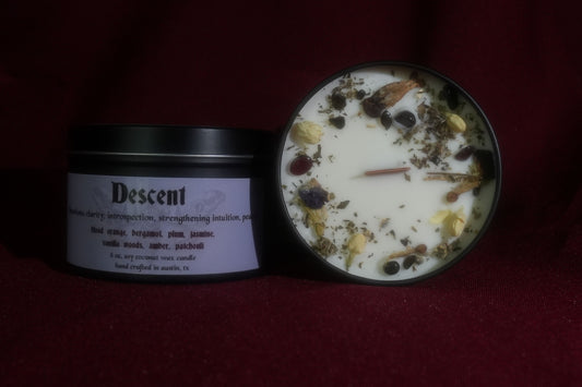 A black metal candle tin labeled 'Descent' sits beside an open matching tin showing white wax topped with dried botanicals and small dark berries, set against a dark red fabric background.