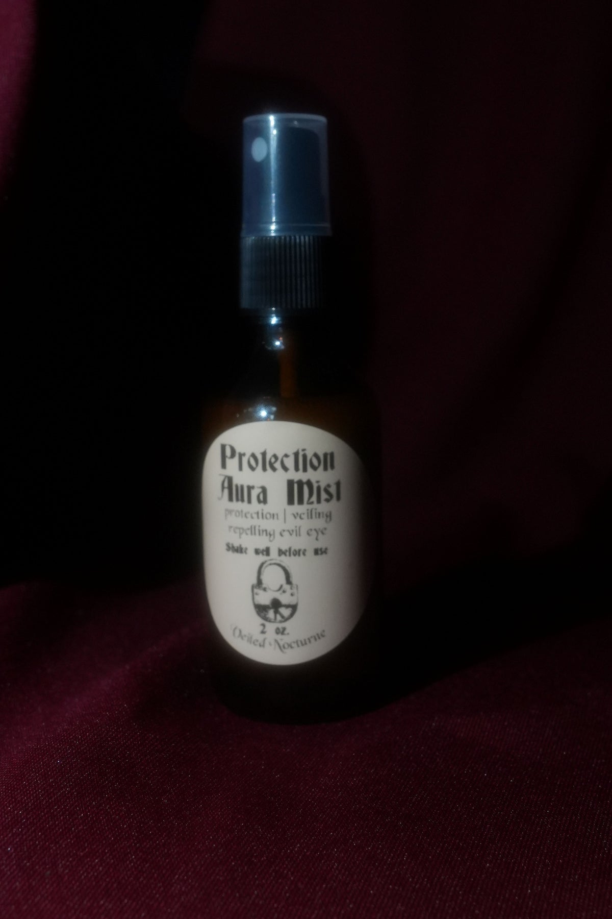 A small amber glass spray bottle labeled 'Protection Aura Mist' with a black spray top and white label, photographed against a dark red fabric background.