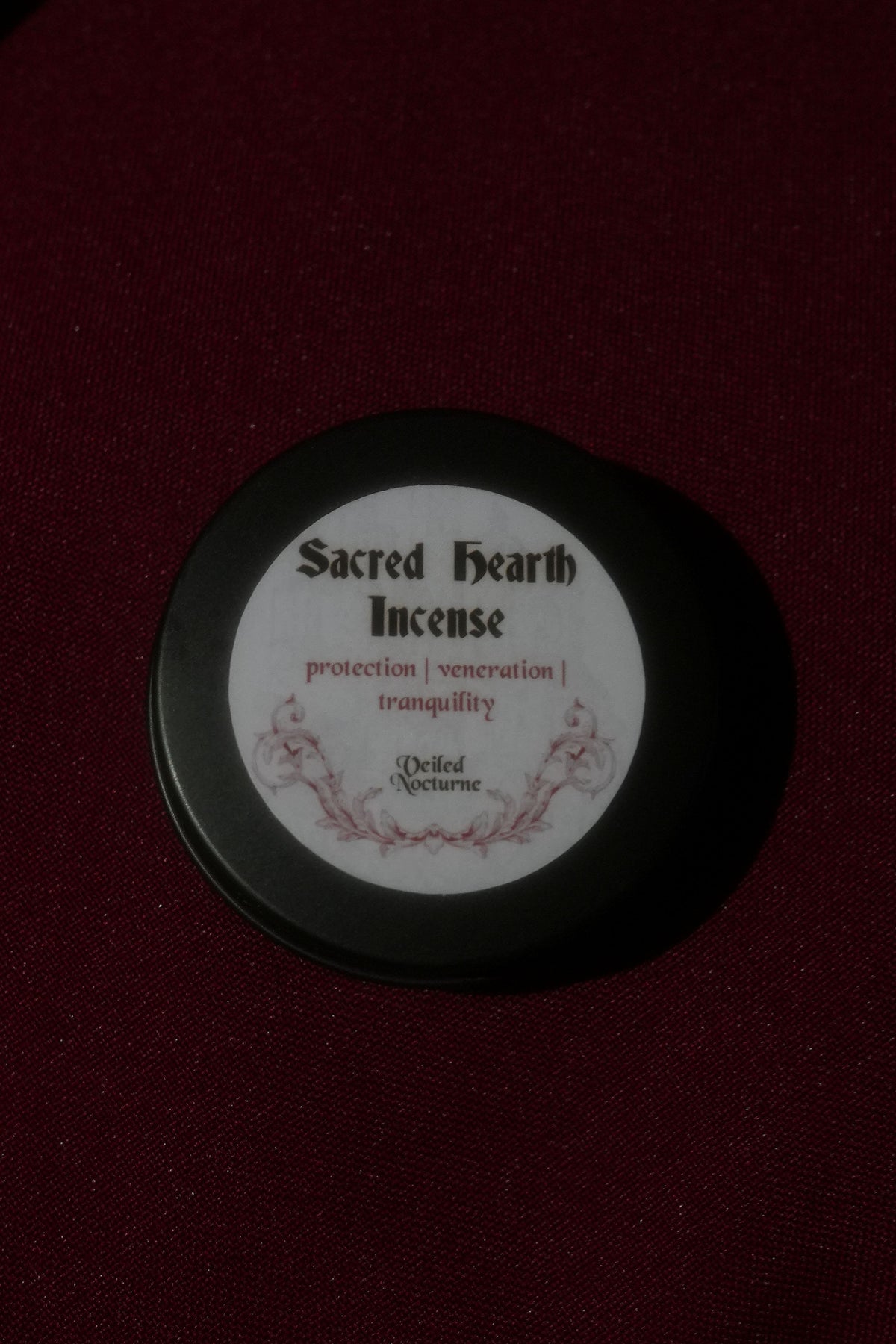 A round black tin of Sacred Ixith Incense on a dark burgundy fabric background, featuring a white circular label with black text and a small red floral illustration.